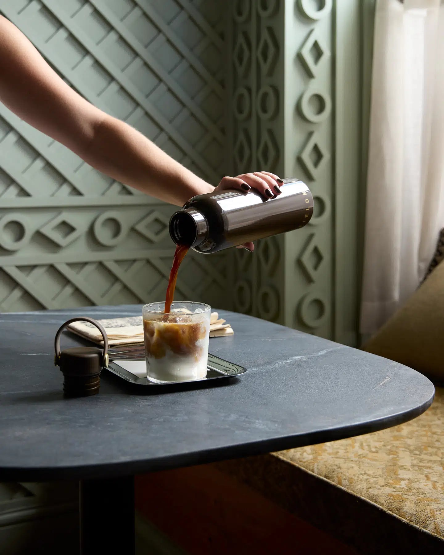 Person pouring coffee from a fieldbar bottle into a glass on a table with a decorative wall in the background.